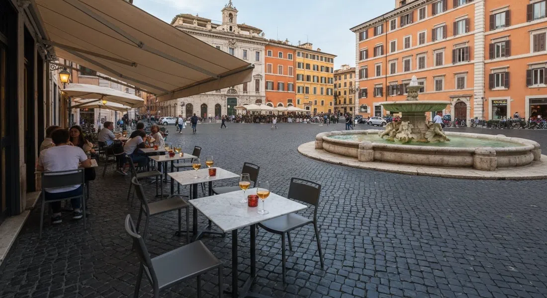 La terraza de La Bottega del Caffè en la Piazza della Madonna dei Monti, asientos sombreados frente a la fuente con copas de spritz nocturno en mesas pequeñas